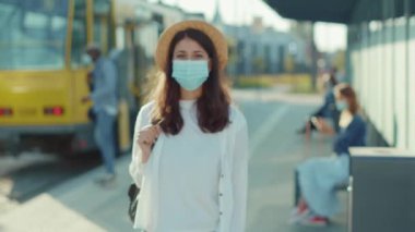 Tıbbi maskeli güzel bir kadını kapatın. Kameraya bakın ve otobüs durağında durun. Pozitif ruh hali olan turistler yaz tatillerinde dinleniyorlar.