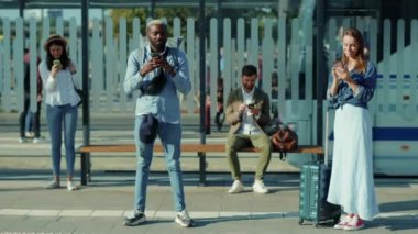 Mutlu karışık ırklar genç erkekler ve kadınlar sosyal mesafelerini koruyorlar otobüs durağında telefon kullanıyorlar Afro-amerikalı tatilciler dışarıda bekliyor ağır çekimde turistler