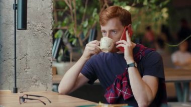 Havalı kafenin içinde cep telefonuyla konuşan kızıl saçlı yakışıklı adam içeride sıcak espresso içiyor. İletişim. İnsanlar ve teknoloji.
