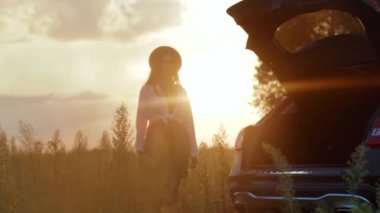 Şapkalı mutlu genç bir kadın akıllı telefon kullanıyor güneş ışığında şık bir arabanın bagajında oturuyor cep telefonu özgürlüğü nakliye aracı kız macera teknolojisinin tadını çıkarıyor yavaş çekim