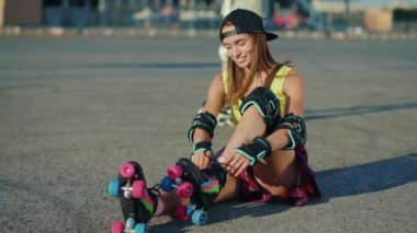 Açık hava stili genç bir kadın yerde oturuyor ve patenler koyuyor gülen sokaklar yaz aylarında bacaklar ağır çekimde patenle kayıyor.
