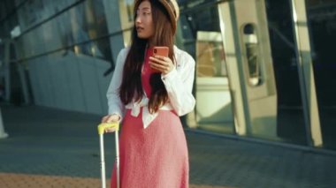 Güzel Asyalı çekici bir kadının iletişim kurmak için akıllı telefonu kullandığı uzun bir çekim. Harika yaz şehrine varmak için havaalanı terminalinin yanında duruyor. Çinli kız. Seyahat.