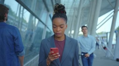 Sosyal medyada haberleşen akıllı telefonlu Afro-Amerikan kadın yönetici. Havaalanı binası. İş merkezi. Kalabalık.