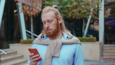 Açık havada gülümseyen ve akıllı telefon tutan yakışıklı beyaz adamın portresi. Caddede cep telefonu kullanan kızıl sakallı yakışıklı adam..