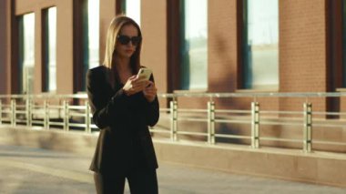 Güneş gözlüklü kendine güvenen iş kadını, iş merkezinin yakınında güneş ışığı giysisi içinde telefonla yürüyor. Başarılı teknolojiyi yavaş çekimde çekici buluyor.