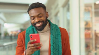 Gülümseyen Afrikalı Amerikalı genç adam Noel zamanı AVM 'de telefon yürüyüşü yapıyor. Noel ağacı mutlu alışveriş yapıyor.