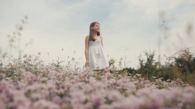 Ahenkli gülümseyen beyaz, romantik elbiseli beyaz kız sahada el kaldırıyor. Bitki, çiçekler. Muhteşem bir manzara. Yaz zamanı. Açık havada. Köy.