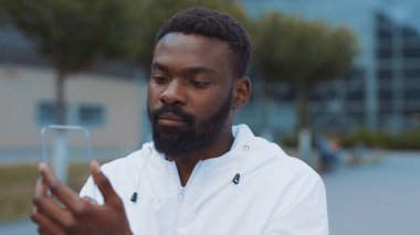 Gülümseyen Afro-Amerikalı adam şehirde fütürist cam telefon yürüyüşü yapıyor. Kameraya bak. Nano-martfon şeffaf elektronik gelecekteki küresel bilgi yenileme arayüzü.