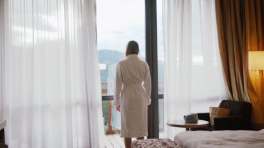 Arkadan bakan genç bir kadın dantel perdeleri açıyor. Lüks bir apartman dairesinde ya da otelde durup pencereden bakıyor. Hafif şehir gökdelenlerinin keyfini çıkarıyor.