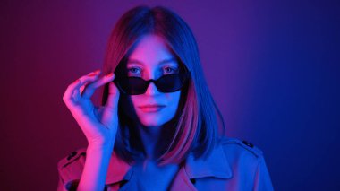 Güneş gözlüklü klas genç bayan kameraya bakıyor, gülümsüyor. Şık giyinmiş güzel bir kızı vurmak. Keyfin yerinde. Neon ışıkları. Mor, renkli. Fikir..