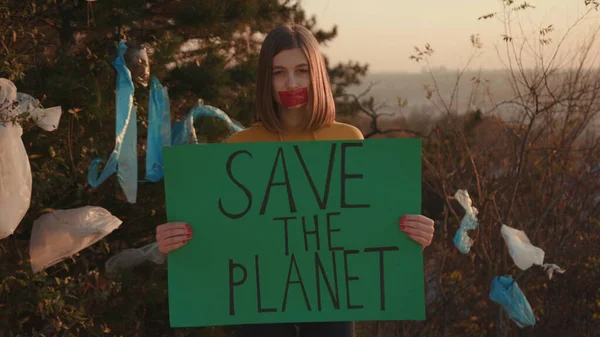 Sad young woman activist mouth glued holding encouraging green poster Save the Planet standing at landfill site with garbage nature environmental community earth eco environment outdoor