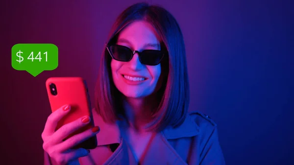 App Icon with Online Transaction. Financial Transactions in the Smartphone. Receive a Message About Increase Money. Attractive Young Woman in Black Sunglasses Holding Red Smartphone in Hand Looking at