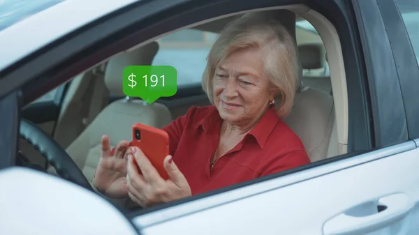 Happy Senior Woman Sits In Car Use Red Cell Phone Smile Businesswoman Blonde. App Icon with Online Transaction. Financial Transactions in the Smartphone. Receive a Message About Increase Money.
