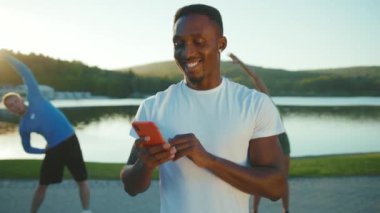 Mutlu Afro-Amerikan fitness eğitmeni akıllı telefon dijital cihazı kullanırken arka planda kaslarını esneten aktif bir çift kardiyo egzersizleri yapıyor..