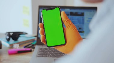 NEW YORK - 28 Mart 2020: Genç bir iş adamı evinde bilgisayarla çalışan dikey krom anahtar modelleme ekranında akıllı telefon kullanıyor. Evdeki insanlar. Teknoloji. Karantina.