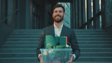 Resmi giyinmiş, gülümseyen mutlu genç adam iş merkezinin önünden geçiyor ve kişisel eşyaları kovulmuş, yavaş çekimde işsiz kalıyor.