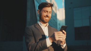 Şehir merkezindeki iş adamı durağında çekim yapmak için telefon mesajlaşması, kaydırma, güler yüzlü gülümseme teknolojisi, güneşli gün başarısı, ağır çekim.