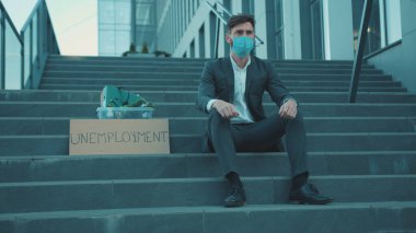 Merdiven başında oturan endişeli genç adam kişisel eşyalarıyla ve yazıtlı posteriyle Coronavirus krizi yüzünden işten atıldı iş dünyası ağır çekim ekonomisi