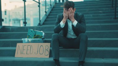 Merdivenlerde elinde kişisel eşyalar ve yazılı posterle oturan hayal kırıklığına uğramış genç adam Coronavirus krizi yüzünden işten atıldı.