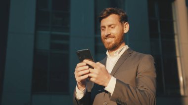 Şehir merkezindeki mutlu işadamı duruşu telefon mesajlaşması, kayan gülümseme teknolojisi, güneşli gün başarısı yavaş çekim kullanıyor.