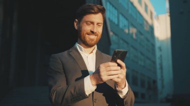 Şehir merkezindeki mutlu işadamı duruşu telefon mesajlaşması, kayan gülümseme teknolojisi, güneşli gün başarısı yavaş çekim kullanıyor.