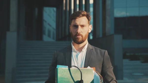 Handsome young man in formal clothes smiling shows middle fingers passing by the business center holds box with personal belongings being fired jobless slow motion