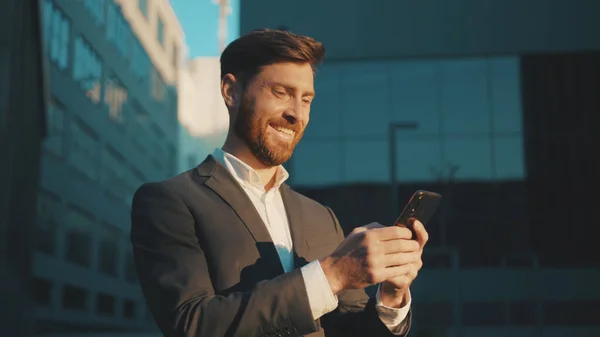 Happy businessman stand in the city center street uses phone texting scrolling tapping smile technology communication sunny day success slow motion