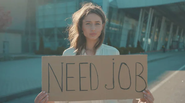 In need of job. Young unhappy woman holding a job search banner hoping for employment striking outside business building. Demonstration. Unemployment, crisis 2020.