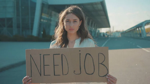 In need of job. Young unhappy woman holding a job search banner hoping for employment striking outside business building. Demonstration. Unemployment, crisis 2020.