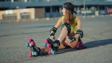 Genç bir kadın yerde oturuyor ve patenleri yerleştiriyor. Elini sallıyor. Gülümsüyor.