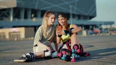 Patenlerle yerde oturan iki genç kadın telefonda gülümsüyor yaz aylarında mutlu bir cadde hissediyorlar bacaklar ağır çekimde patenle kayıyorlar