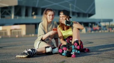 Patenlerle yerde oturan komik genç kadınlar telefon kullanır. Gülümserler. Yaz aylarında mutlu bir cadde hissederler.