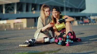 Gün ışığında patenlerle yerde oturan iki genç kadın telefon kullanıyor gülümsüyor ve yaz aylarında mutlu bir cadde hissediyorlar bacaklar ağır çekimde patenle kayıyorlar.