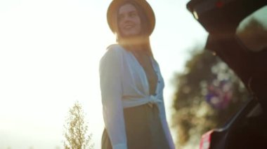 Şapka tezgahı olan genç bir kadın arabasının yanına bakar ve gün batımını seyreder.