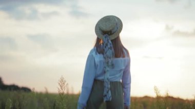 Gün batımında, şapkalı çekici bir kadın kameraya doğru dönüyor. Sıcak doğa tatilleri. Yavaş çekimde genç portre.