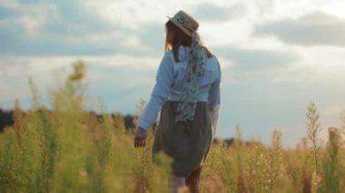 Sahada, güneş ışığında yürürken şapkalı ve elbiseli bir kadın. Sıcak doğa tatili. Genç portresi yavaş çekimde kapanıyor.