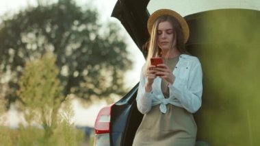 Şapkalı genç bir kadın güneş ışığında şık bir araba bagajında akıllı telefon kullanıyor. Sahada cep telefonu, fotoğraf özgürlüğü, turizm, otomobil, araba macera teknolojisinin tadını çıkarıyor.