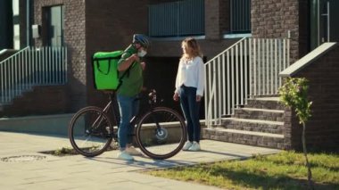 Genç adam kuryesini güvenlik kaskı ve tıbbi maskeyle kapatın. Müşteriye bisiklet çekici bir kadınla kese kağıdıyla yemek dağıtın. Dışarıda durun.