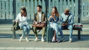 Çok ırklı genç gezginler otobüs durağında bankta oturuyor ve konuşuyorlar. Sokağın ortasında, güneşli yaz aylarında, açık havada ağır çekimde turistler kullanılıyor.