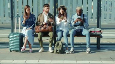 Otobüs durağında çok ırklı genç gezginler bankta oturup konuşuyorlar. Birlikte telefon kullanıyorlar. Yazın şık, güneşli, açık hava turistleri.