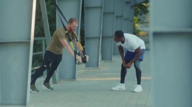 NEW YORK - 5 Eylül 2020: Afrika kökenli Amerikalı genç adam, spor giyim sporlarının yavaş olduğu stadyum kaslı spor giyim antrenmanlarında trx fitness halatıyla dışarıda vücut egzersizleri yapan birini motive etti