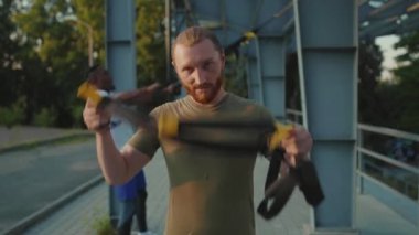 Gülümseyen yakışıklı genç adam, trx fitness halatıyla geçiş yaptıktan sonra kameraya bakıyor. Stadyumdaki spor kıyafetleri spor salonunda vücut egzersizleri yapıyor. Yavaş çekimde spor portresi çekiliyor.