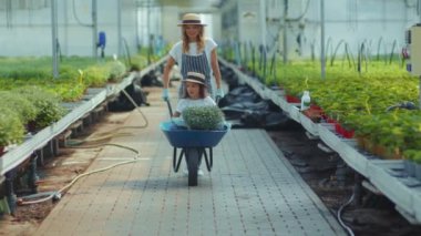 El arabası çiçekli komik bir kızı olan genç bir anne yaz yeşili ailede serada kameraya gülümsüyor kadın çocuk doğası açık hava çocukluğu ağır çekim