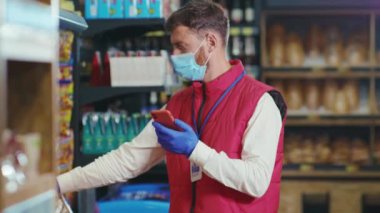Maskeli genç satıcı süpermarkette telefon kullanıyor. Karantina zamanı sebze işinde biber tutuyor. Güzel, taze ürünler, perakende satış elemanı yemekleri.