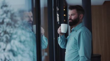 Portre genç yakışıklı sakallı, kahve içen ve pencereden manzaranın tadını çıkaran yakışıklı, erkek çay penceresi dairesi çekici, beyaz, insan yüzü dinlendirici, kapalı mekanda yetişkin panoraması.