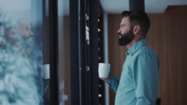 Yaklaş yakışıklı sakallı, kahve içen ve pencereden manzaranın keyfini çıkaran yakışıklı, erkek çay penceresi dairesi yakışıklı, beyaz, insan yüzü dinlendirici yetişkin panorama yavaş çekim.