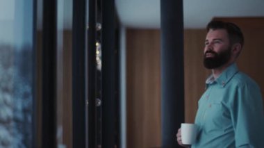 Genç yakışıklı sakallı, mutlu, kahve içen ve pencereden manzaranın tadını çıkaran yakışıklı, erkek çay penceresi dairesi yakışıklı, beyaz, insan yüzü dinlendirici, kapalı mekanda yetişkin panoraması yavaş çekim.