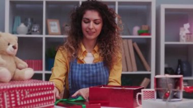 Noel ve yeni yıl dekor paketinin yanında kırmızı kraft kağıdına sarılmış sevimli genç bir kadın. Renkli sürpriz bir gülümseme. Mutlu masa hazırlama portresi.