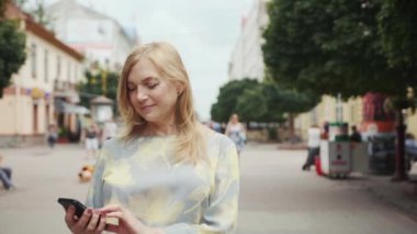 Gülümseyen kadın şehir merkezinde telefon yürüyüşü yapıyor. Mutlu bir yaz interneti. Teknoloji dünyası.