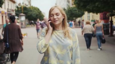 Orta yaşlı mutlu sarışın kadın telefonda konuşuyor. Şehir merkezinde yürürken modern akıllı telefon teknolojisi çalışıyor. İletişim meşgul.